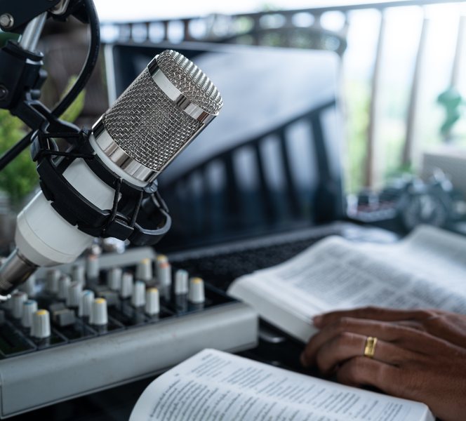 A studio microphone beside a mixing desk, with open books and a hand resting on them